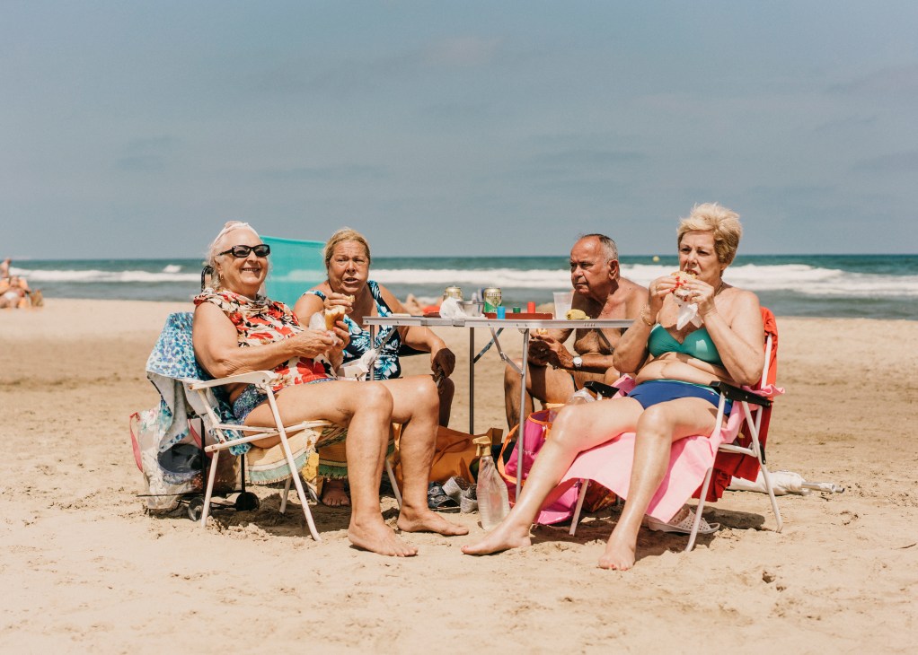 Four seniors enjoy a beach picnic at a table on the sand with the ocean behind them.
