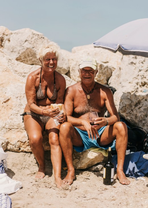 Two older beachgoers sit on sand with white chalk cliffs behind them, sharing food and wine under a beach umbrella.
