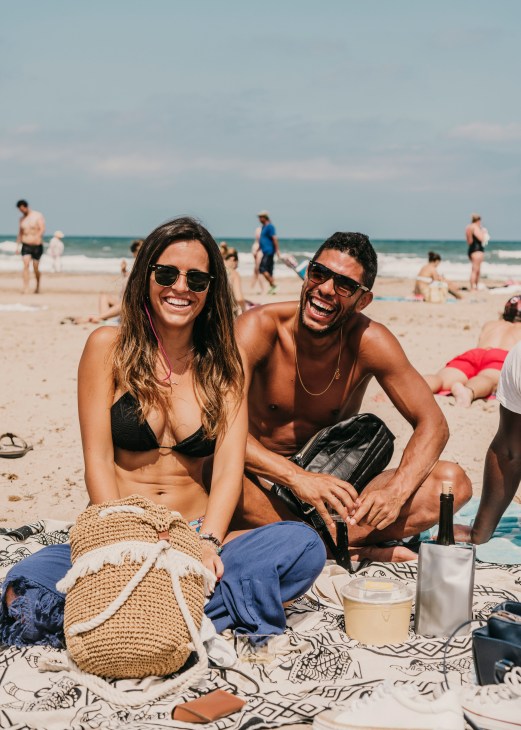 Two people sitting on a patterned beach towel with drinks and a woven bag, smiling at Patacona Beach in Valencia.