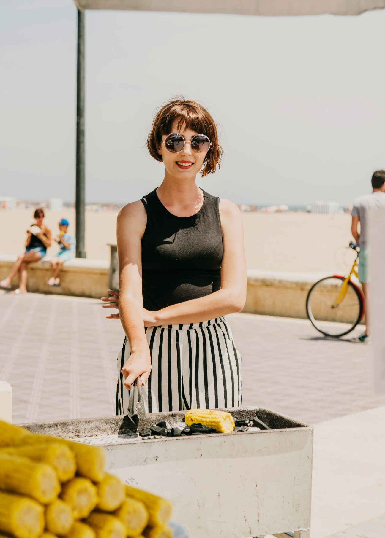 Woman in black tank top and striped skirt with round sunglasses stands at a beach food stall with corn on the cob.