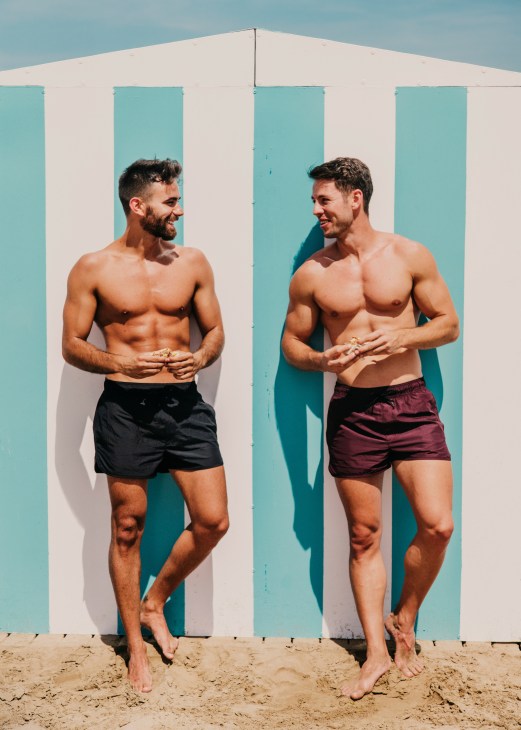 Two shirtless men stand on a beach in front of a turquoise and white striped structure, smiling at each other.