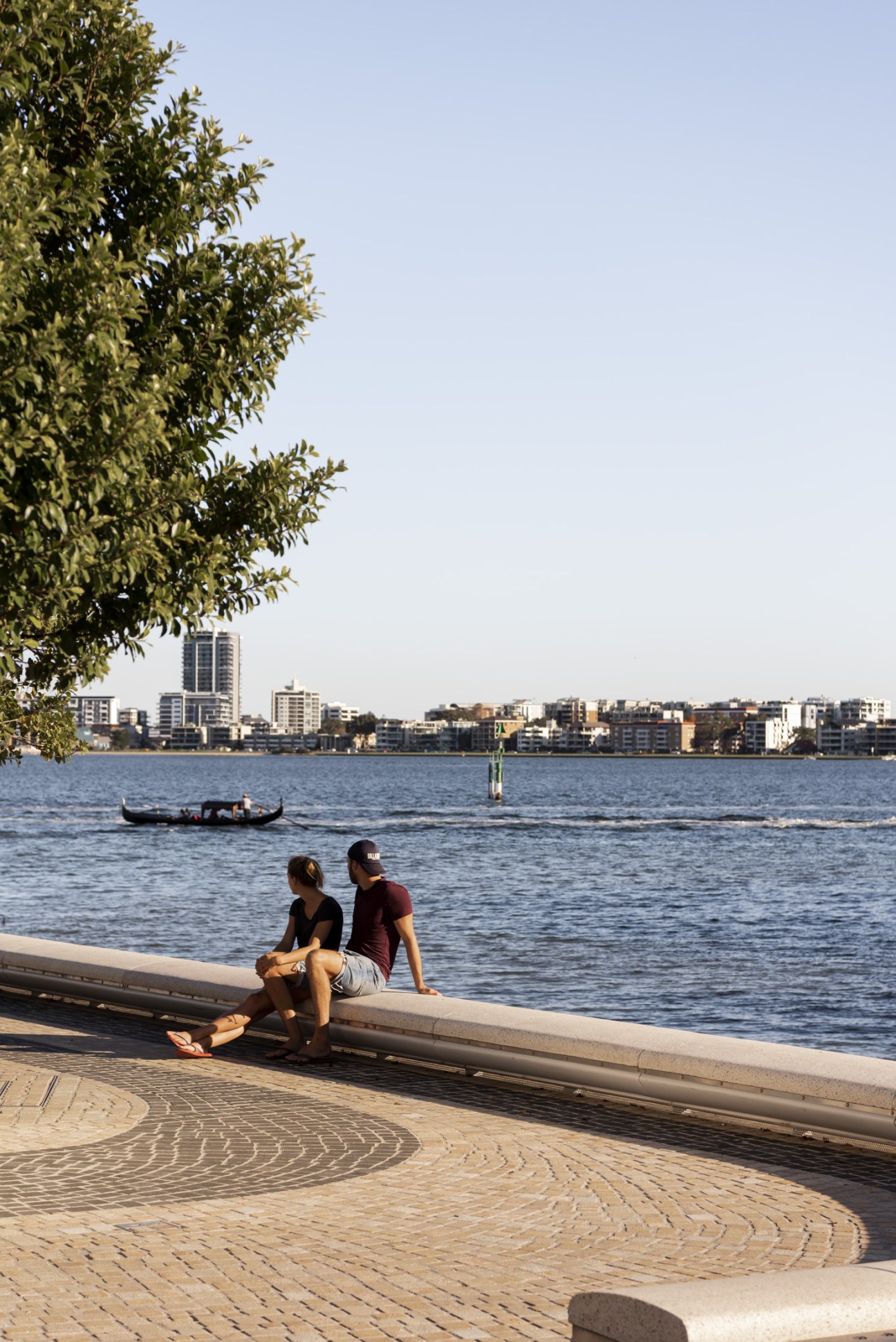 Two people sit on a waterfront promenade at Elizabeth Quay overlooking the Swan River with Perth's skyline and a gondola visible across the water.