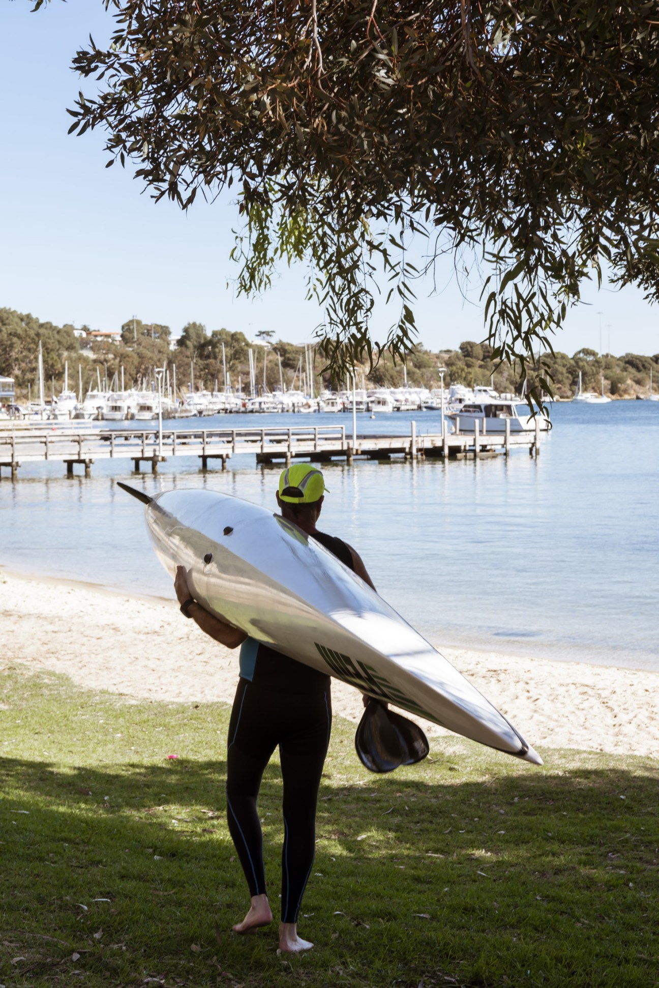 Kayaker in neon helmet carrying boat on Swan River beach at Bicton Baths with moored yachts behind.
