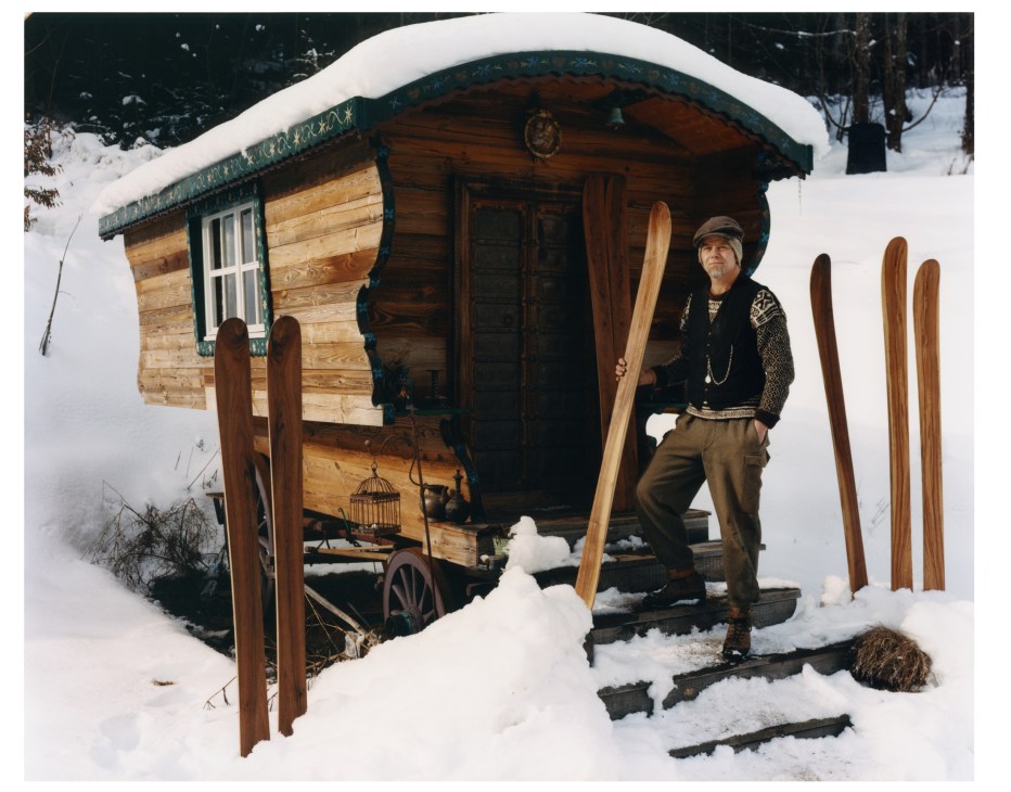 Peter Steltzner stands outside a wooden alpine hut in snow, holding Rabbit on the Roof skis.