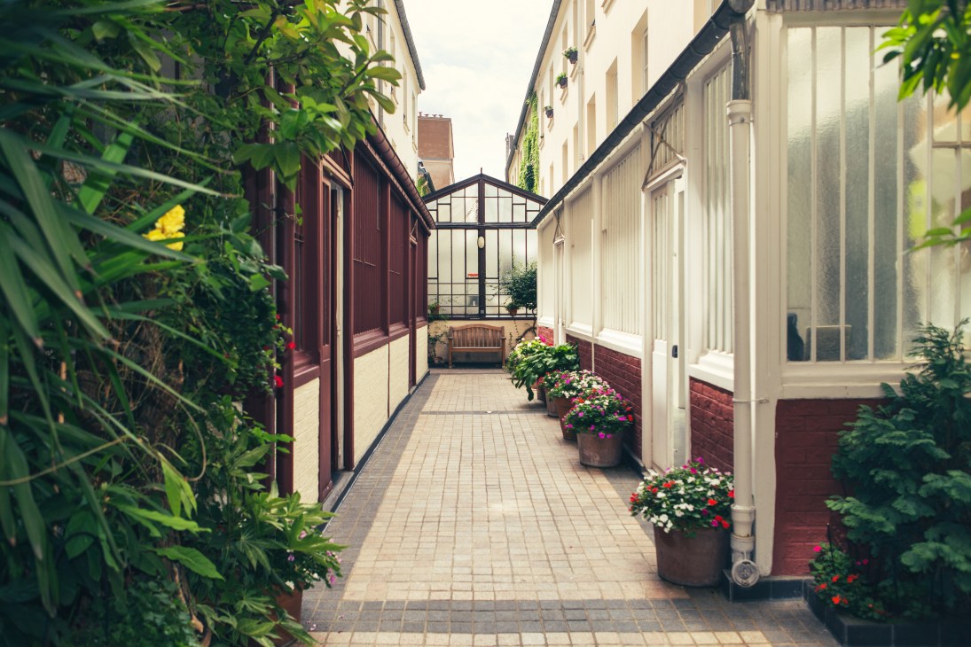 Narrow cobblestone courtyard between cream and red buildings with potted flowers and glass-roofed passage