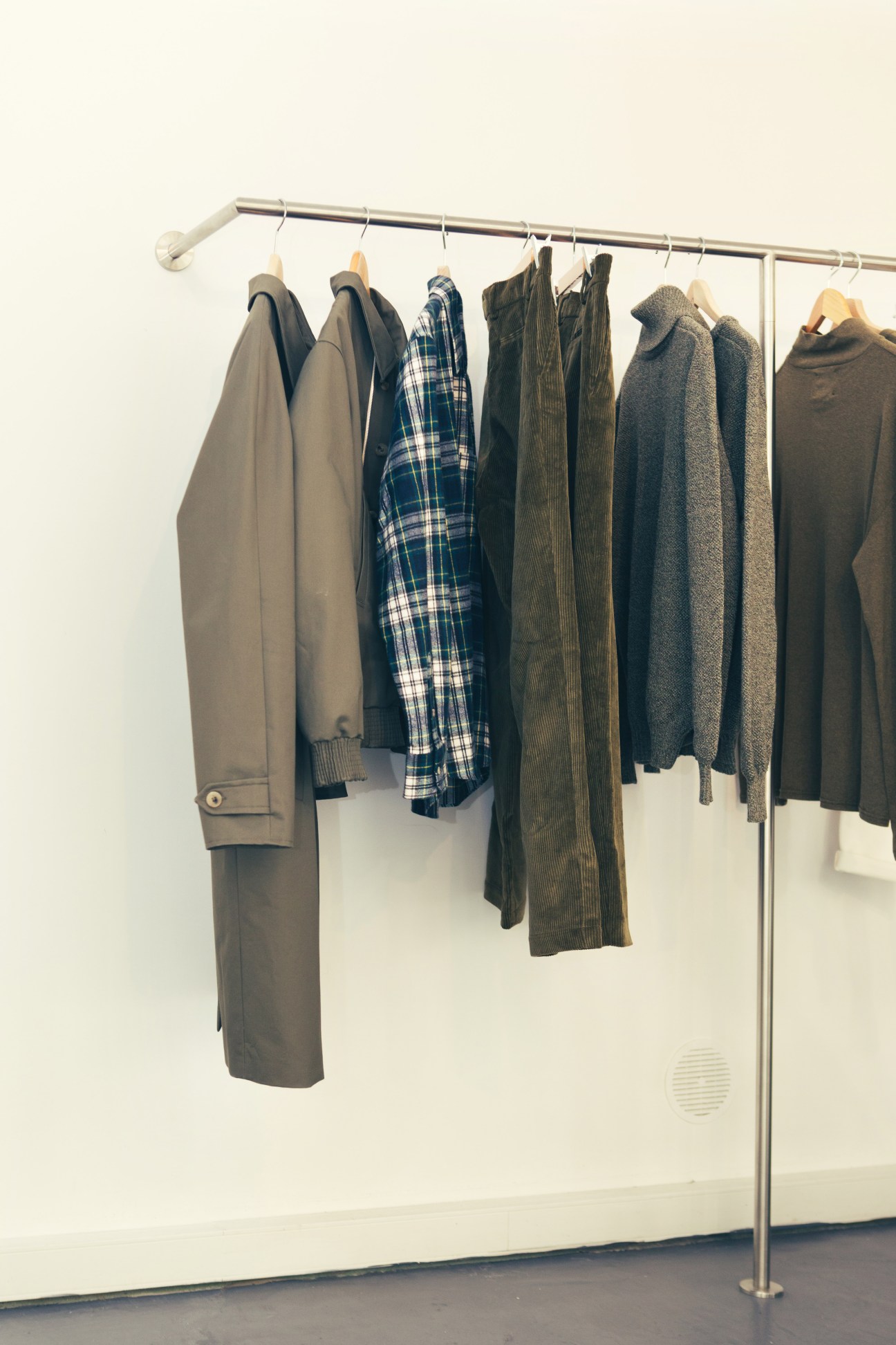 Men's clothing hanging on a metal clothing rack including coats, shirts, and knitwear in muted tones