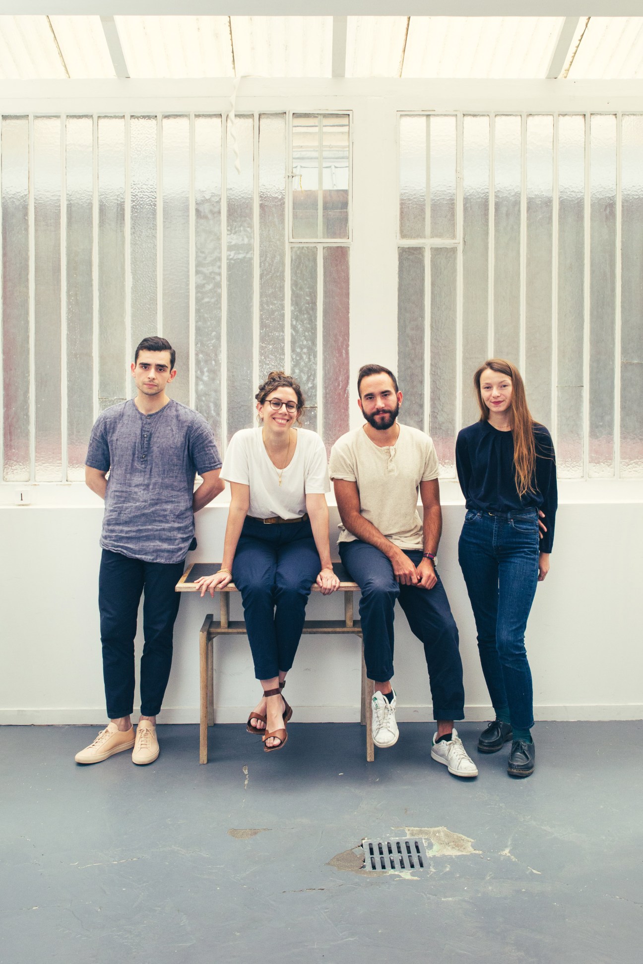 Four people posing together, two standing and two sitting on a bench against a white wall with vertical blinds