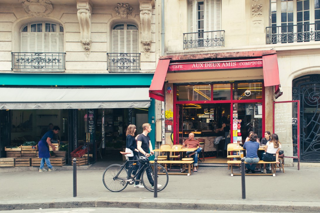 Street view of Aux Deux Amis bistro with red awning, outdoor seating, and people including cyclists on a Parisian street