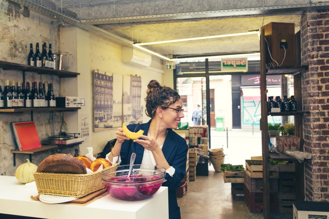 Woman with curly hair and glasses eating banana in grocery store with produce and shelving visible