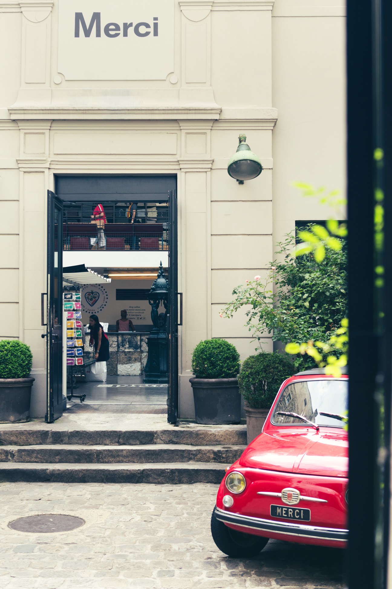 Storefront of Merci concept store in Paris with ornate stone facade, open entrance, and red vintage Fiat 500 parked outside