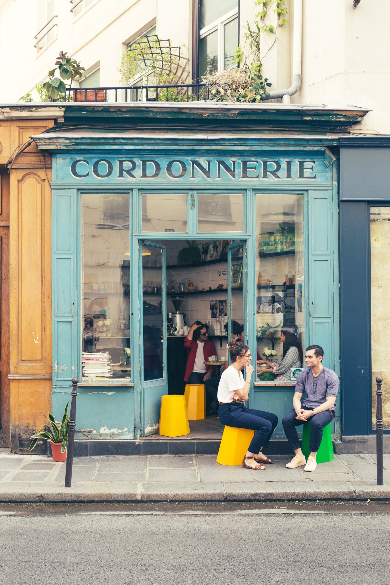 Turquoise vintage storefront of Cordonnerie shoe shop with people sitting on colorful stools outside