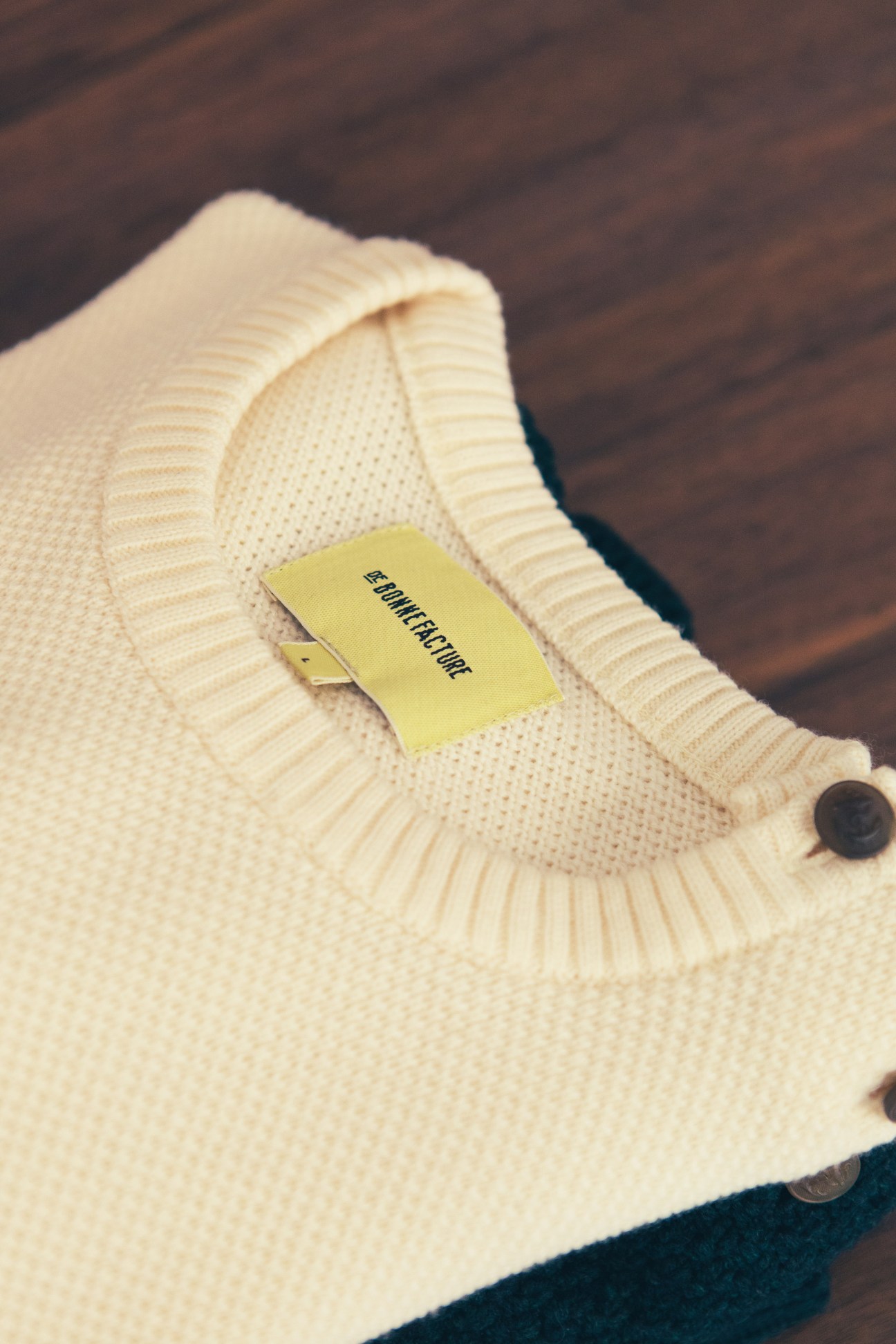 Cream knit cardigan with yellow De Bonne Facture label laid on wooden surface