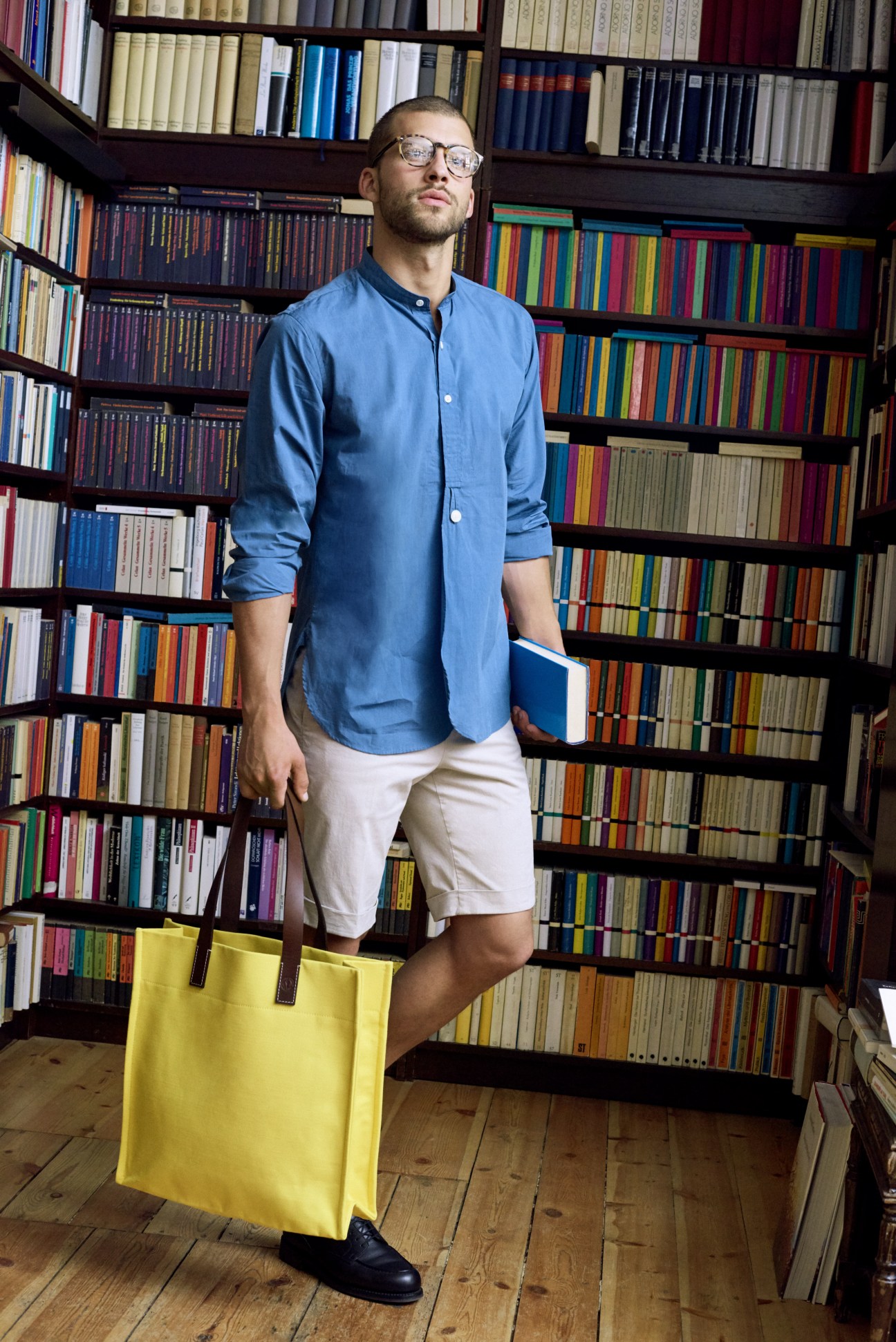 Man in blue shirt and white shorts holding blue book stands in front of colorful library shelves.