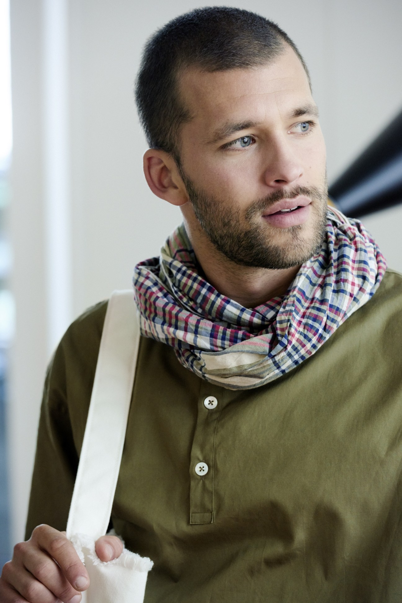 Man wearing olive pullover shirt, checkered scarf and white bag looking to the side.