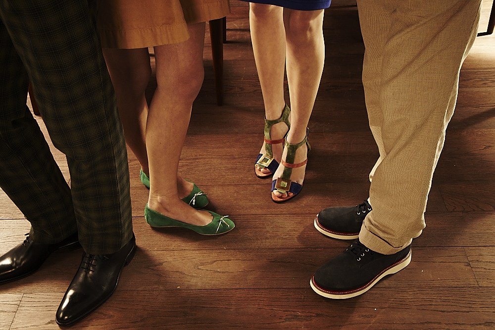 Multiple people's feet and lower legs displayed on a wooden floor, wearing various designer shoes, trousers and skirts.