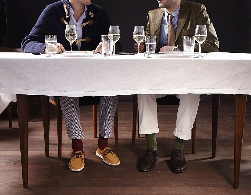 Two men seated at a dining table displaying their lower legs and footwear under a white tablecloth.