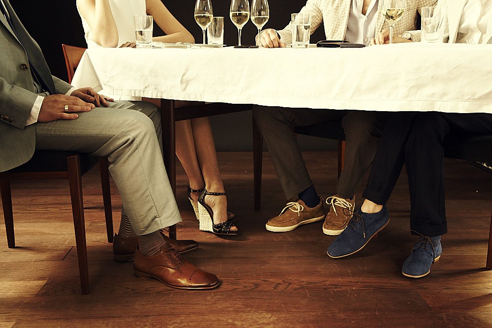 People seated under a dining table wearing designer shoes and trousers, including Brioni, Falke, Camper, Incotex Red and Tod's.