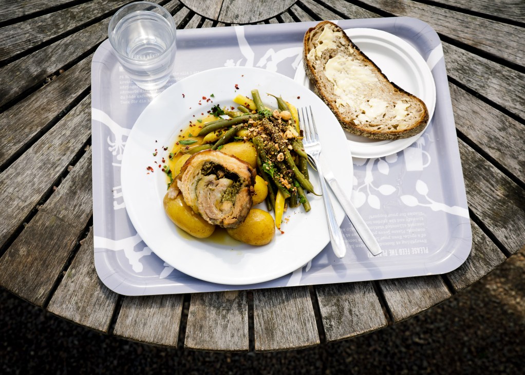 Sliced porchetta with green beans and roasted potatoes on white plate with bread and water glass