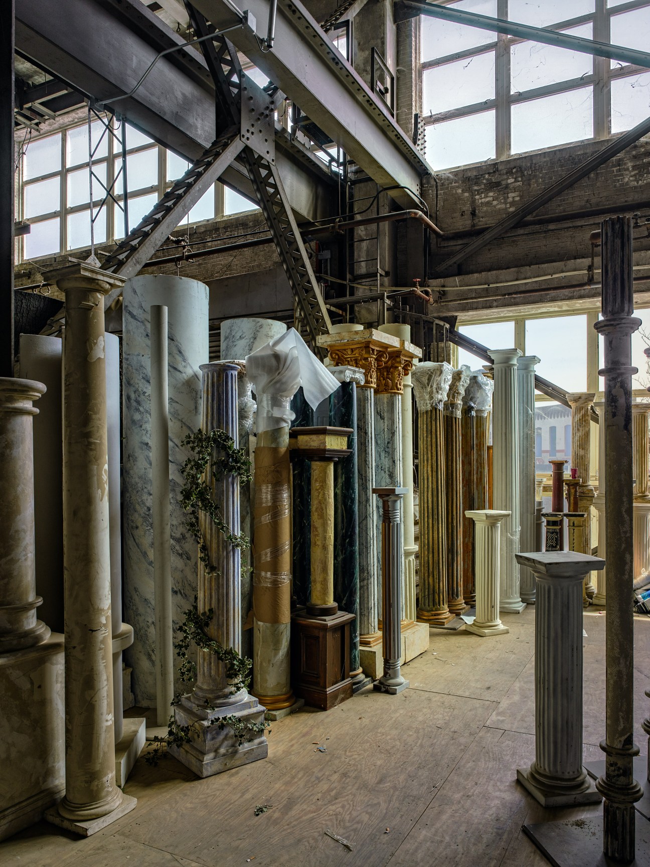 Assorted decorative columns and architectural elements stored in an industrial warehouse space with exposed beams and large windows.