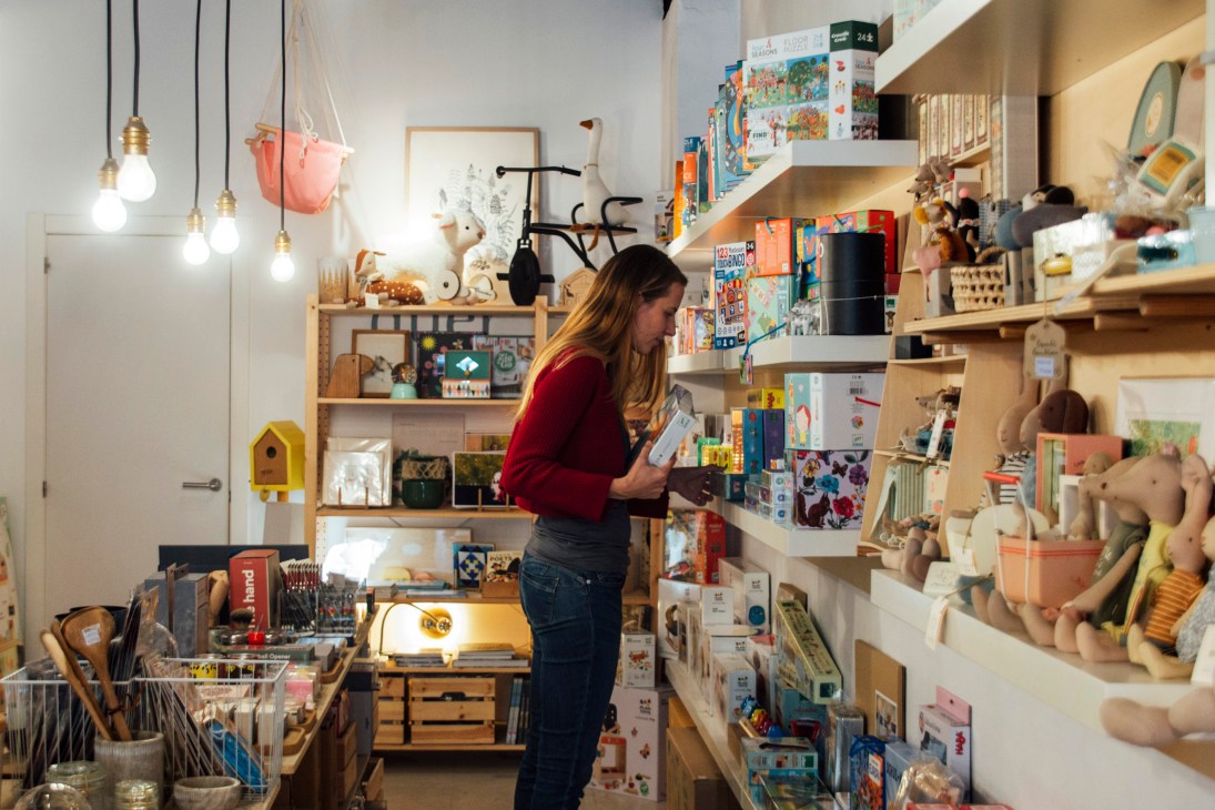 Woman browsing in a bright toy shop with wooden shelves displaying children's toys and books.