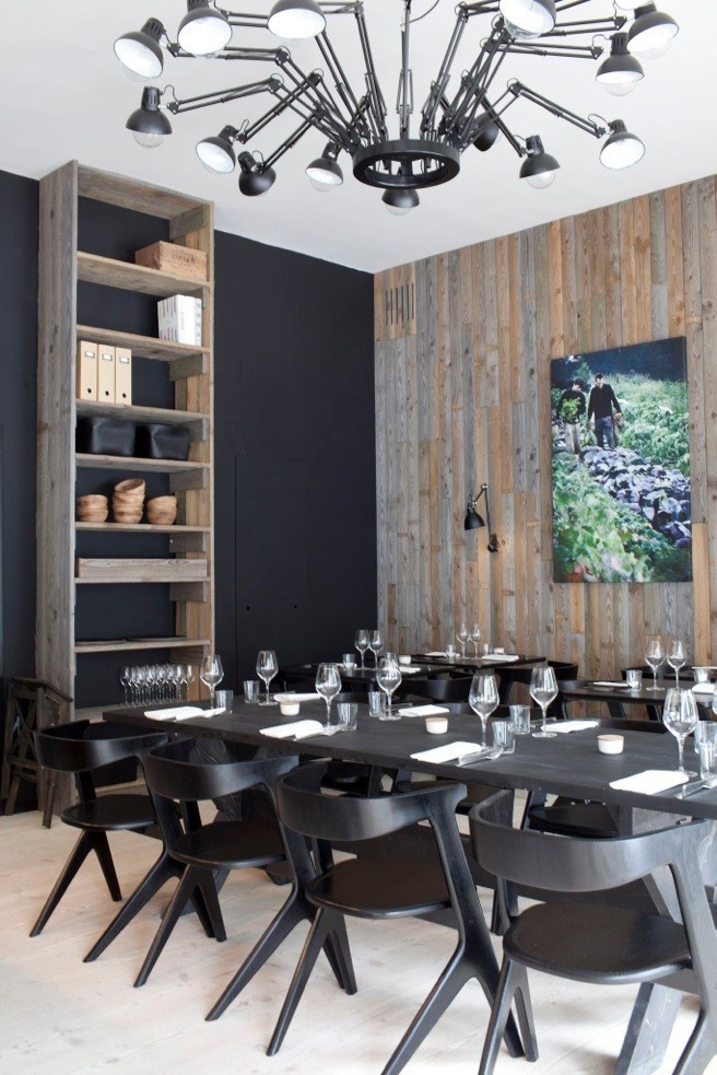 Modern restaurant dining room with black table, chairs, wooden wall paneling and industrial ceiling lights.