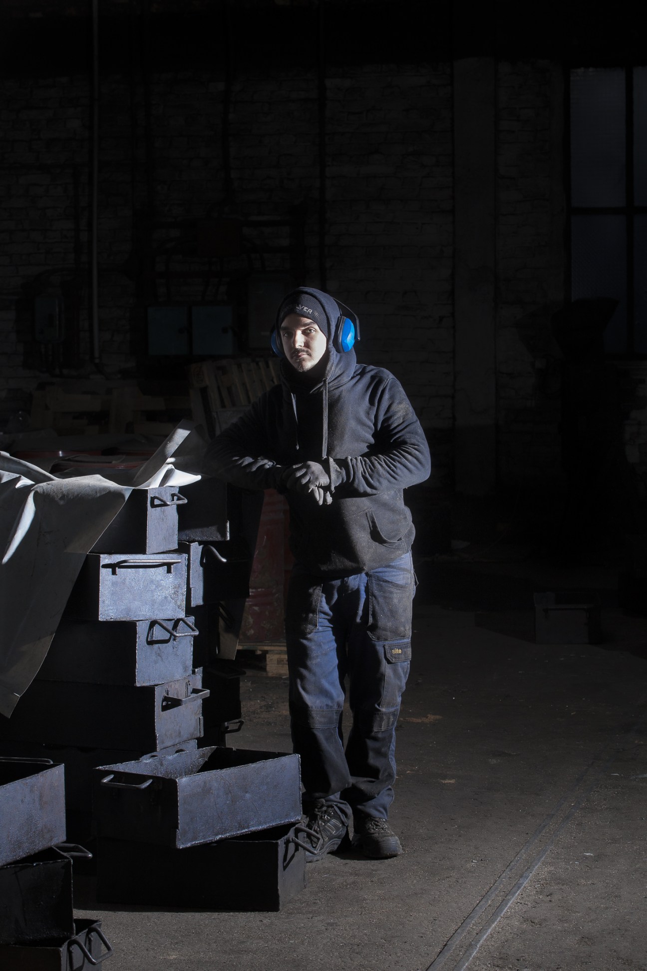 Factory worker wearing headphones and dark clothing stands beside vintage nail-making machinery on the factory floor.