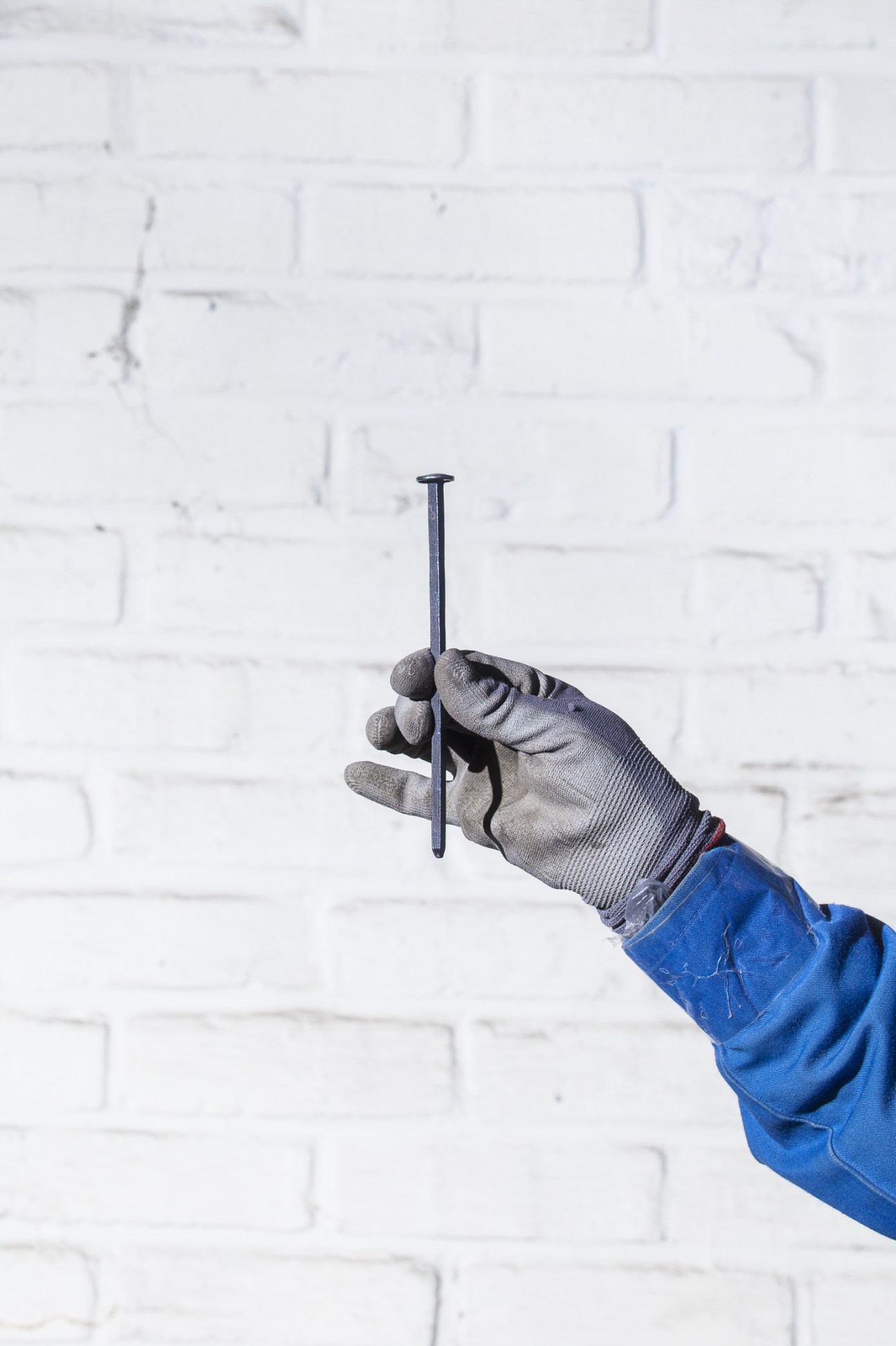 A worker's gloved hand holds a large dark nail against a white brick wall.
