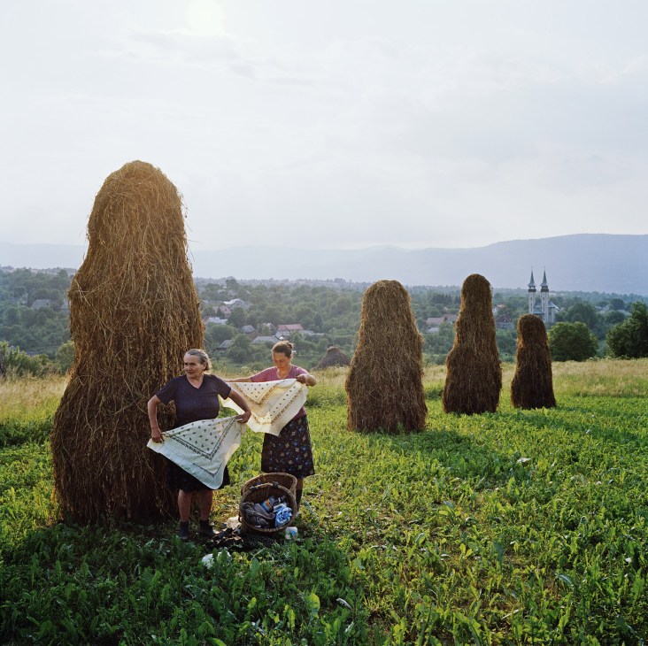 Two women holding fabric near tall hay stacks in a rural Carpathian mountain landscape with a church visible in the distance.