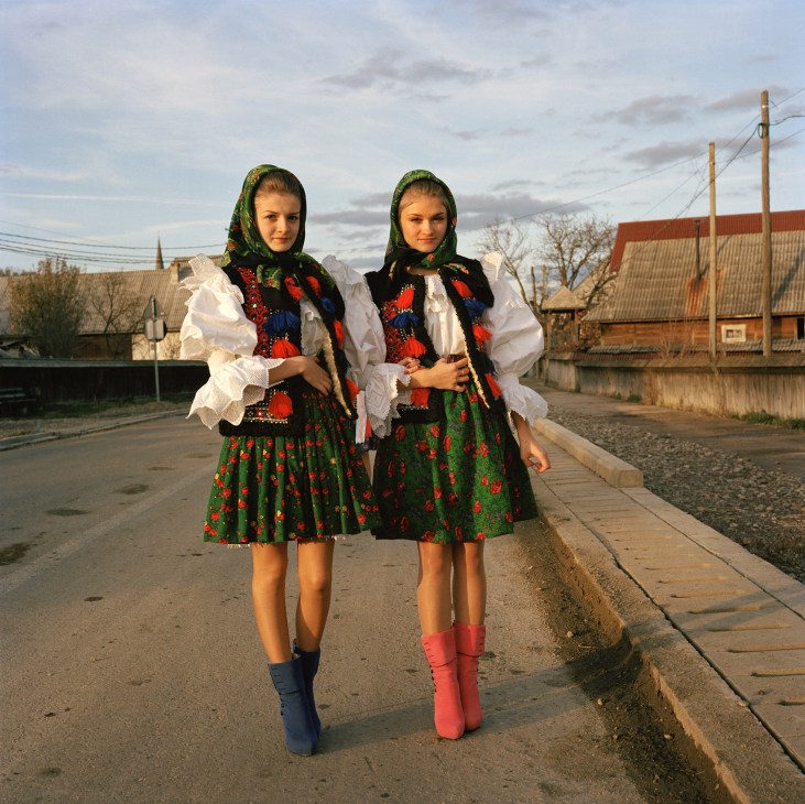 Two young girls in traditional Romanian folk costumes with embroidered dresses and headscarves walk down a rural road.