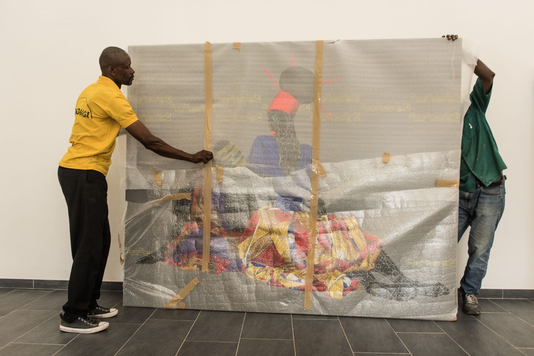 Two men unwrap a packaged painting depicting a Congolese figure wearing red and blue clothing.