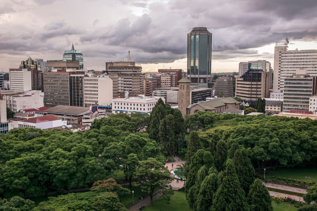 Downtown Harare skyline with colonial and modern buildings under cloudy sky, surrounded by green vegetation.