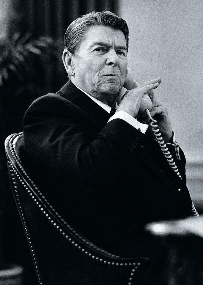 Ronald Reagan sitting in chair with hand to chin, speaking on telephone in black and white photograph.