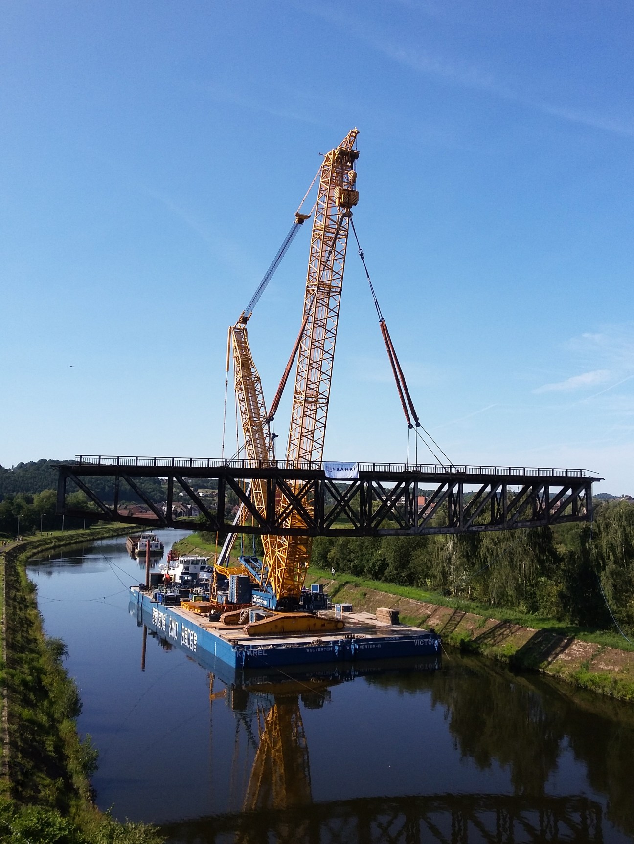 Large yellow crane mounted on a blue barge lifting a steel bridge structure over a river.