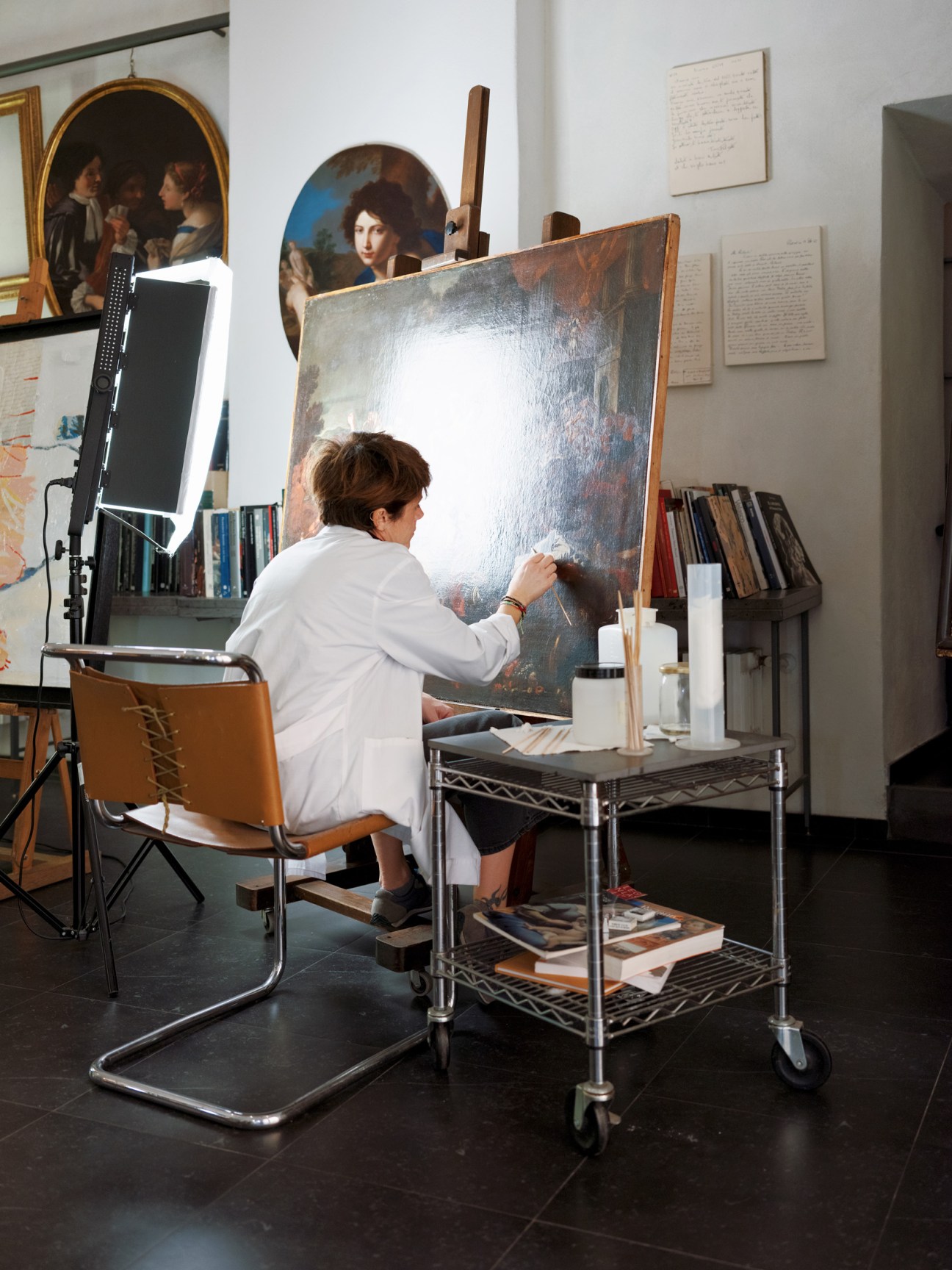 Art restorer in white coat working on a large old master painting on an easel in a studio.