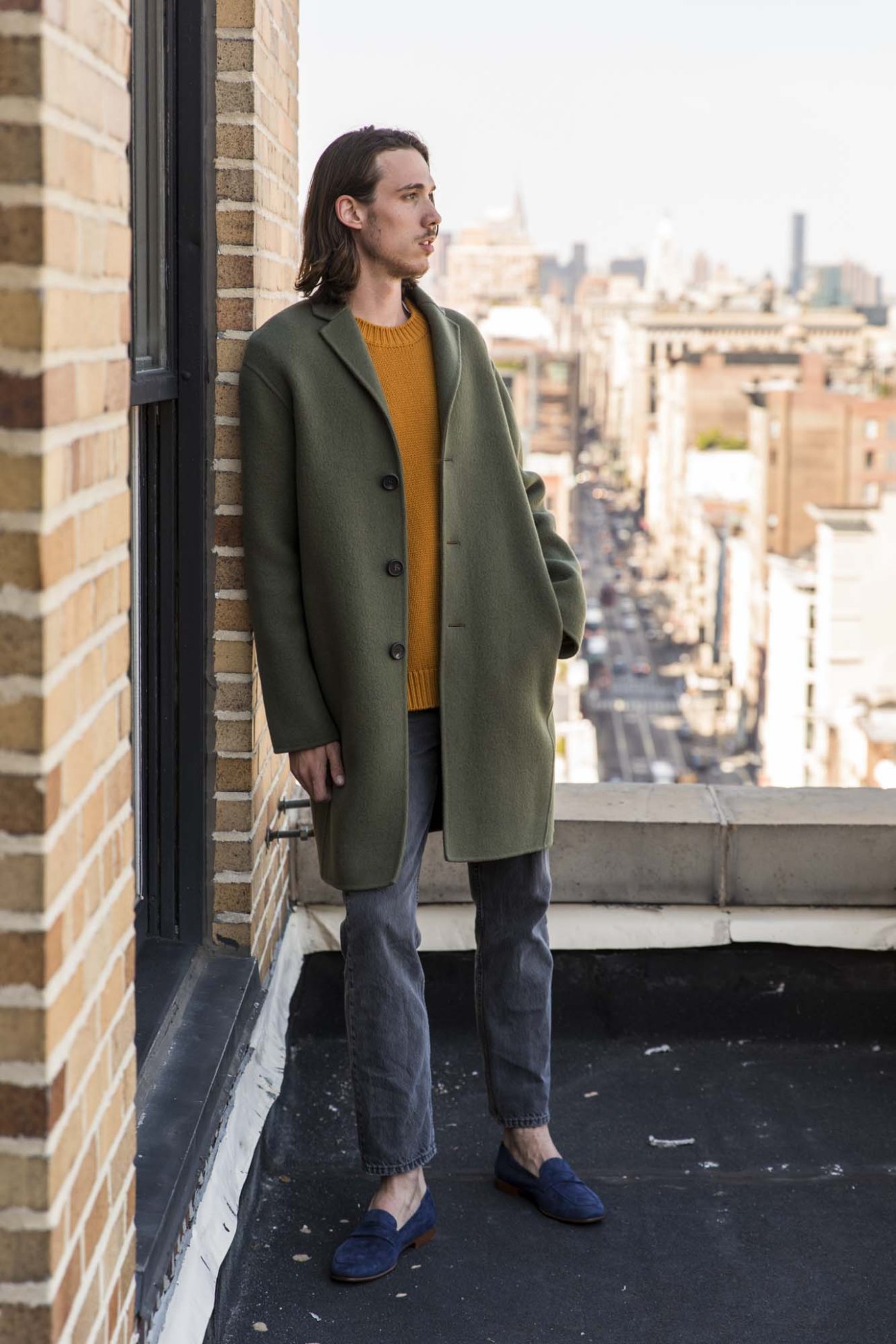 Man wearing sage green boiled-wool coat, mustard cashmere crewneck and navy suede loafers on rooftop overlooking NYC