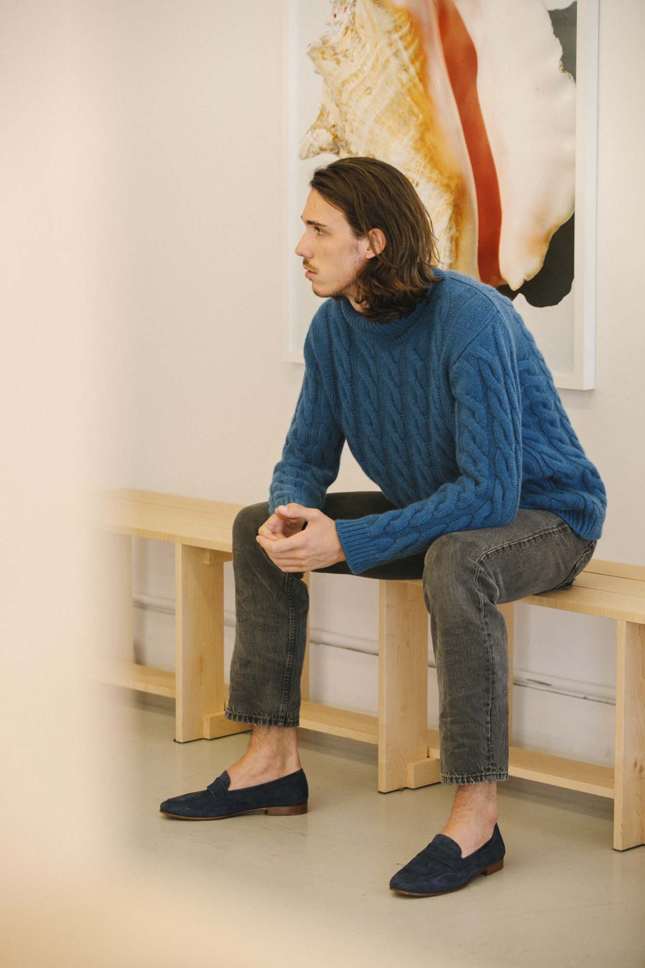 Man wearing blue cashmere cable-knit turtleneck, gray jeans, and navy suede loafers sitting on wooden bench.