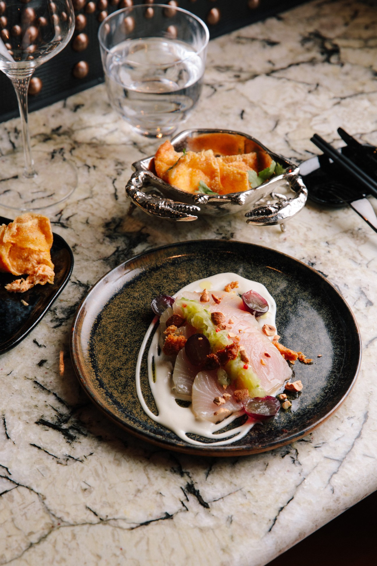 Plated hamachi crudo and snow-crab rangoon at Wild Ink restaurant in Hudson Yards, served on marble table with wine glasses.