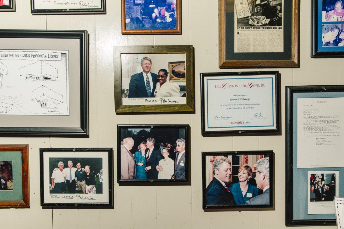 Framed photographs and memorabilia of Bill Clinton displayed on a cream-colored wall at Doe's Eat Place restaurant.
