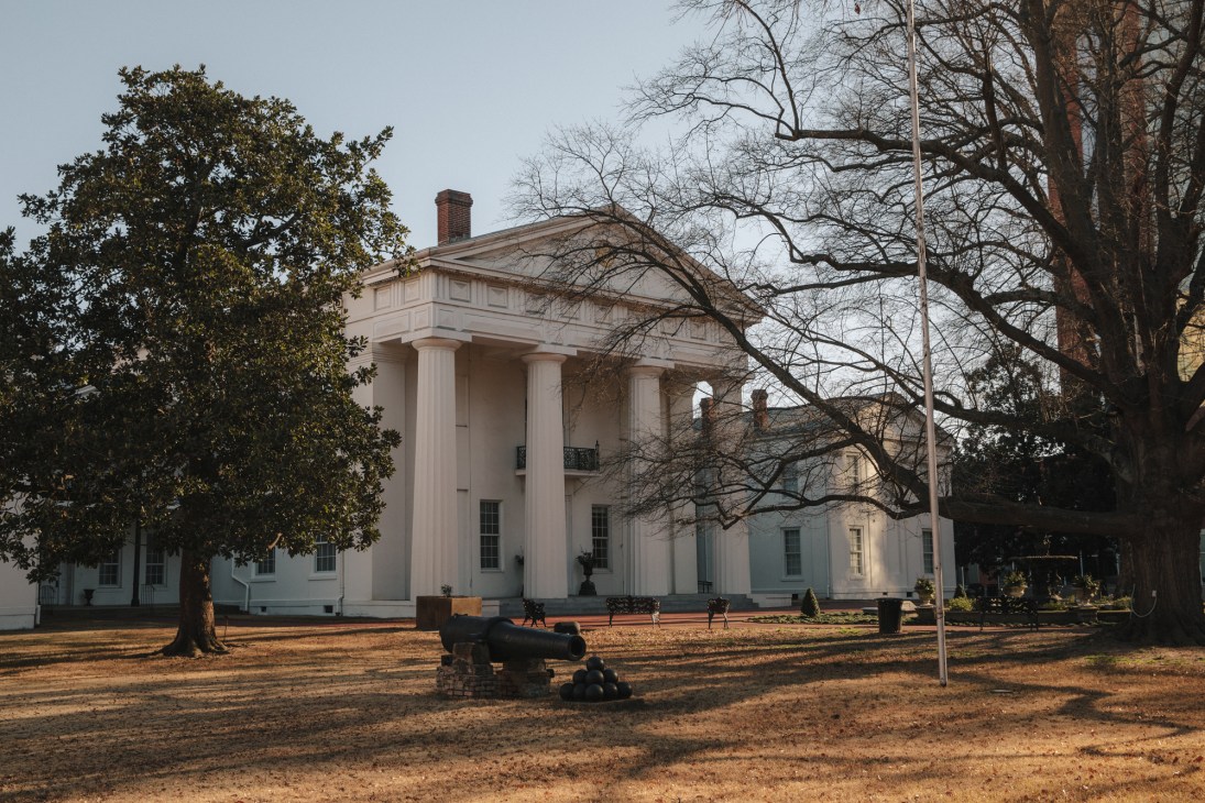 White neoclassical building with columns and brick chimney surrounded by bare winter trees and manicured grounds.