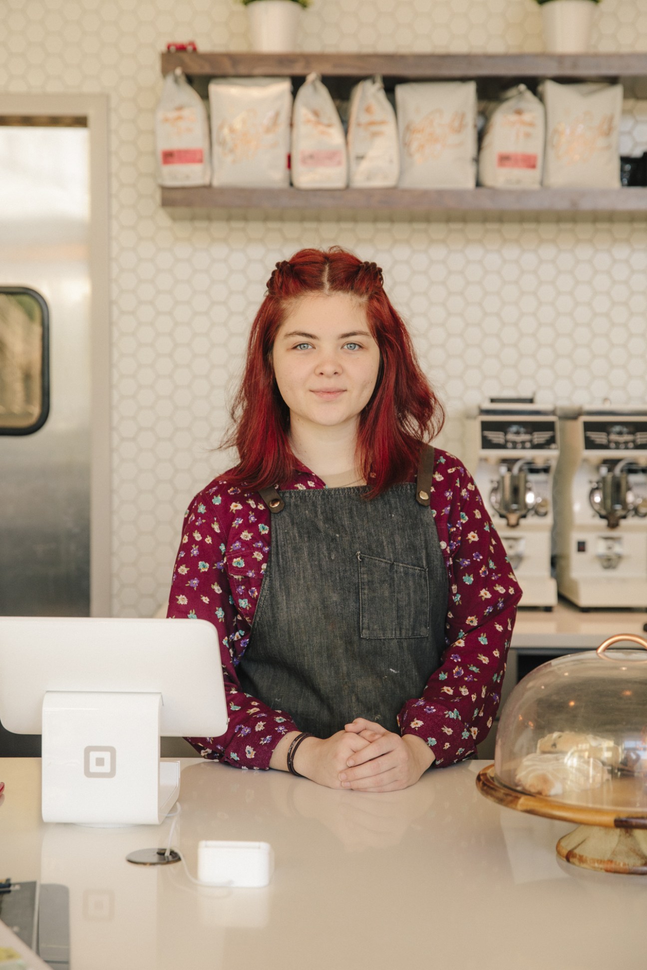 Annabelle, a barista at Zetêo Coffee, stands behind the counter wearing a burgundy floral shirt and denim apron.