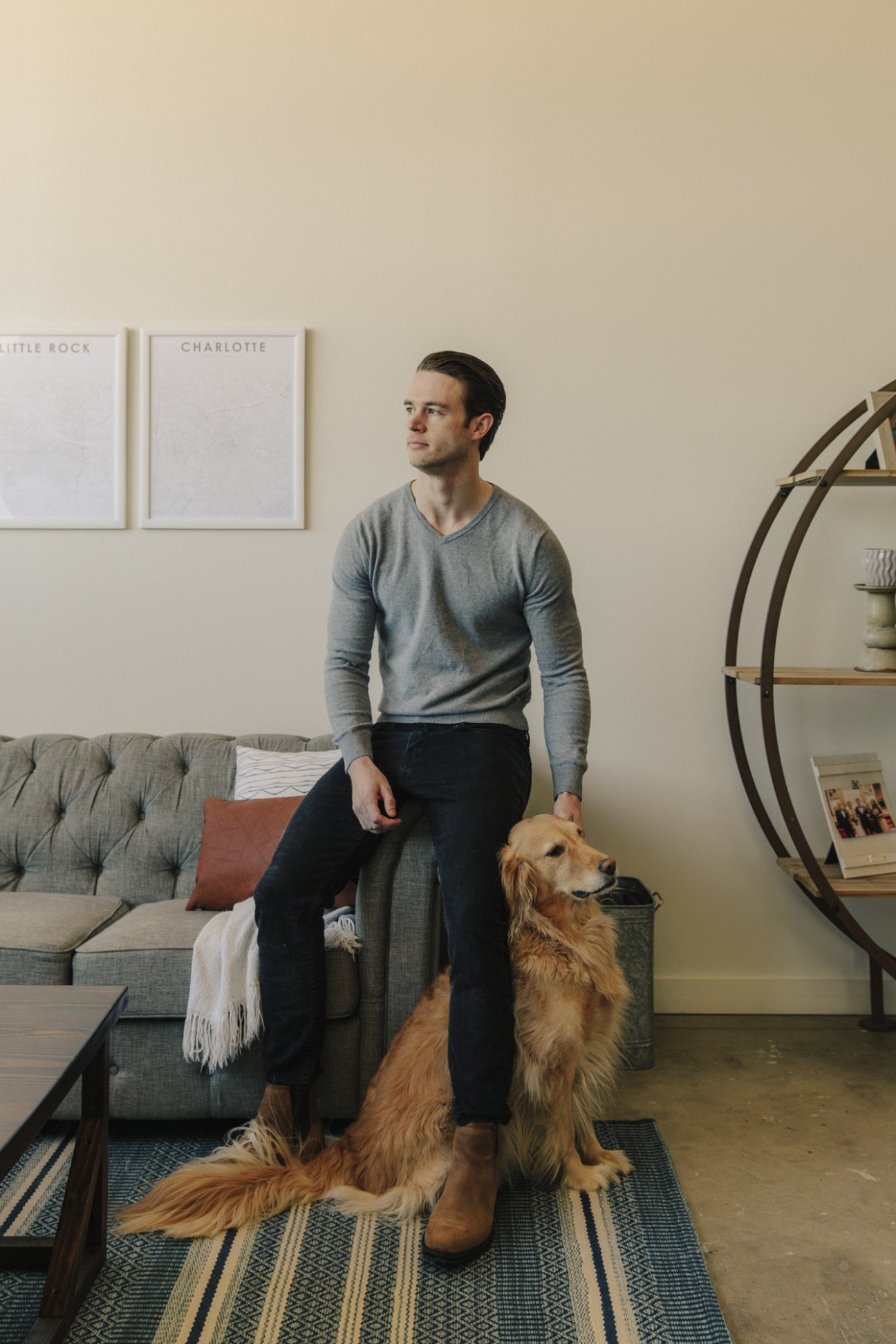 Man sitting on gray sofa with golden retriever, framed city maps on wall behind.