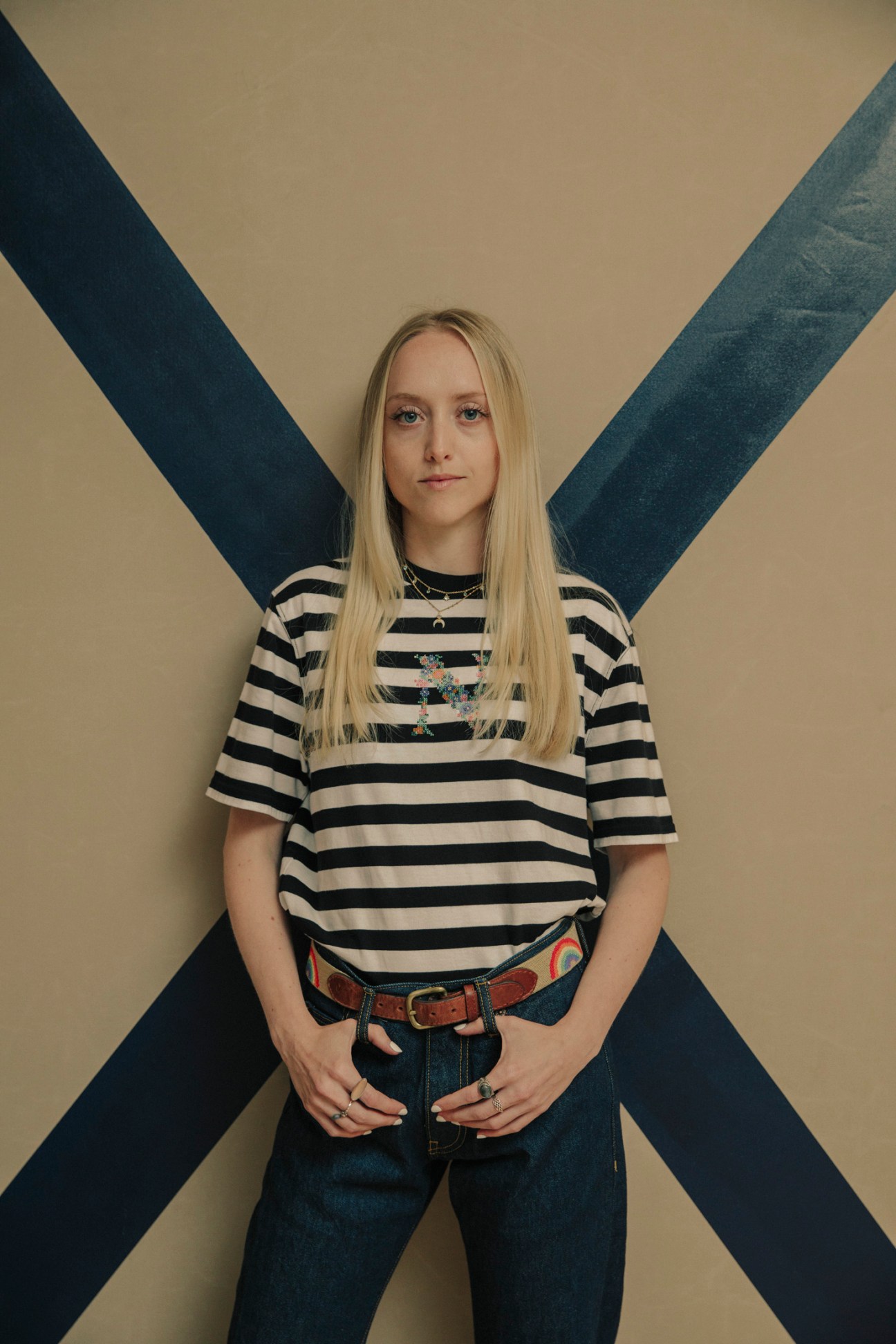Young woman wearing Noah striped top with five-pocket denim and rainbow belt against geometric background.