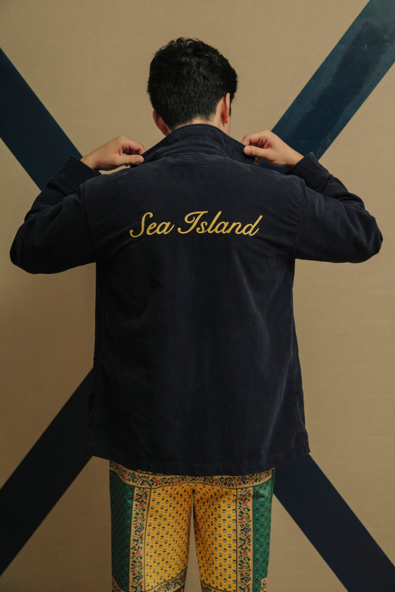 Person wearing black Sea Island jacket with gold embroidered text over patterned yellow and green pants against beige background.