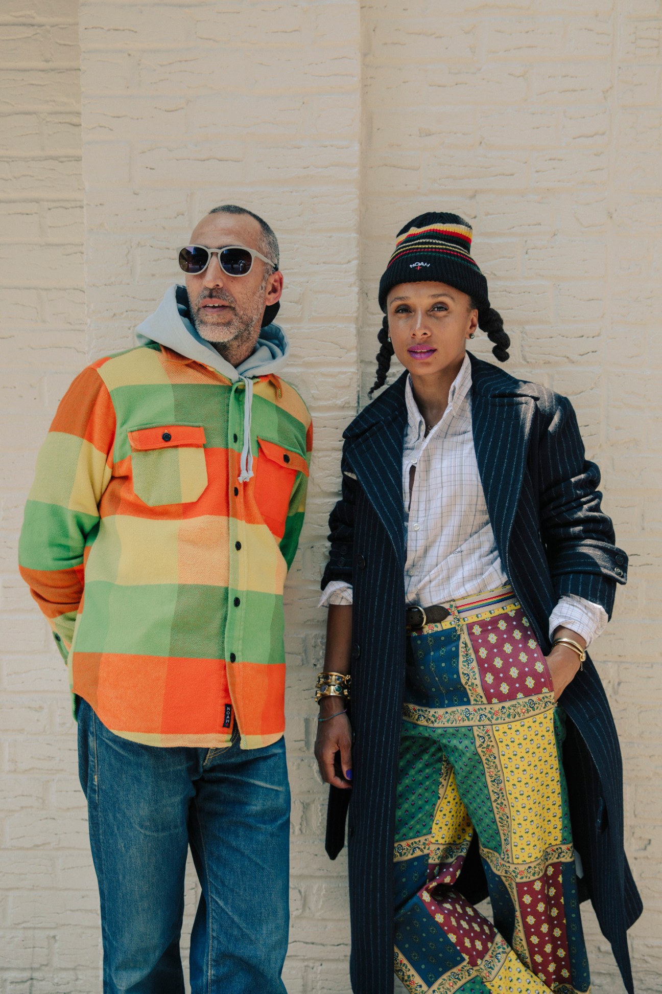 Noah founders Brendon Babenzien and Estelle Bailey-Babenzien pose against a white brick wall wearing colorful clothing from their collection.