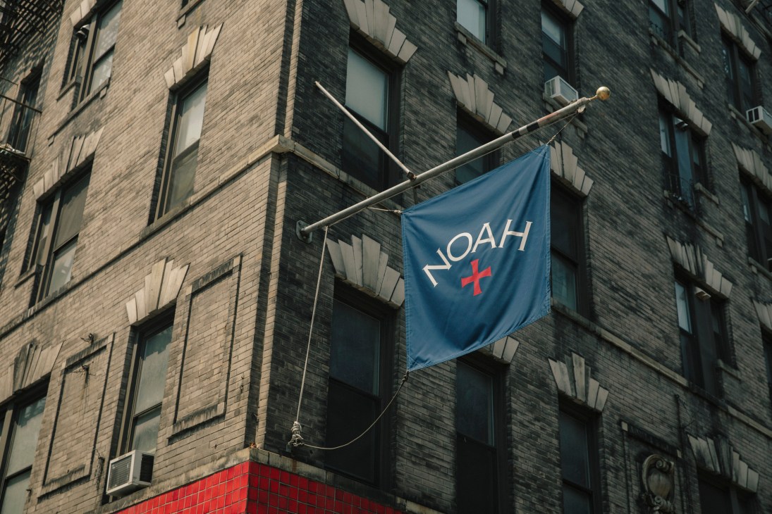 Blue banner with NOAH logo and red plus sign hanging from brick building exterior.
