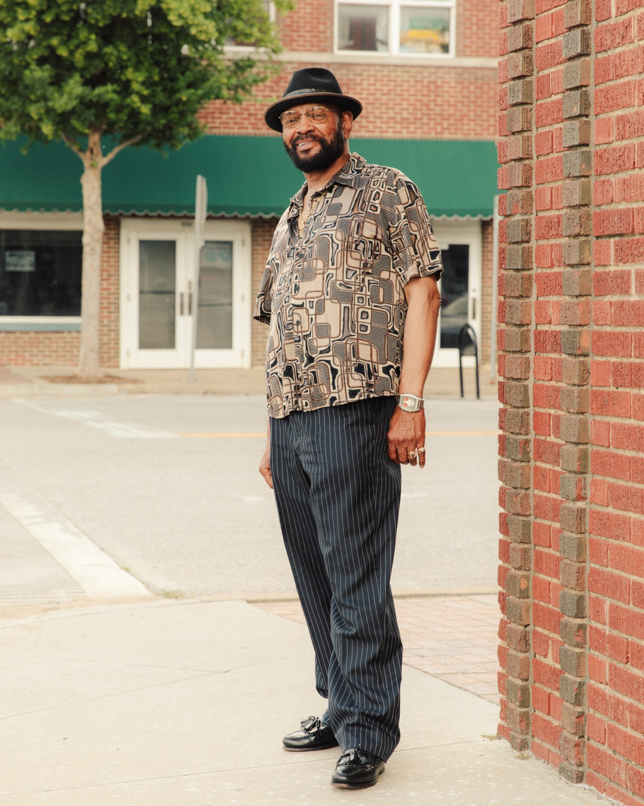Man standing on a brick-paved street wearing a patterned shirt, pinstriped pants, and black hat.