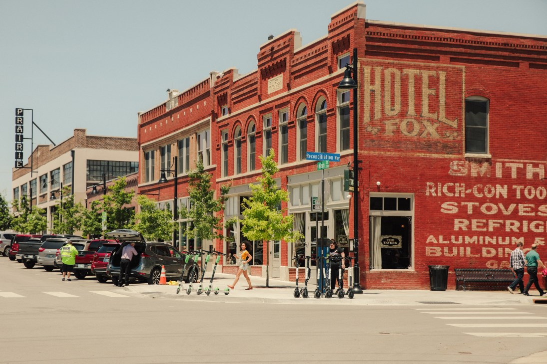 Historic red brick buildings line a street in Tulsa's Arts District with shops, restaurants, and pedestrians.