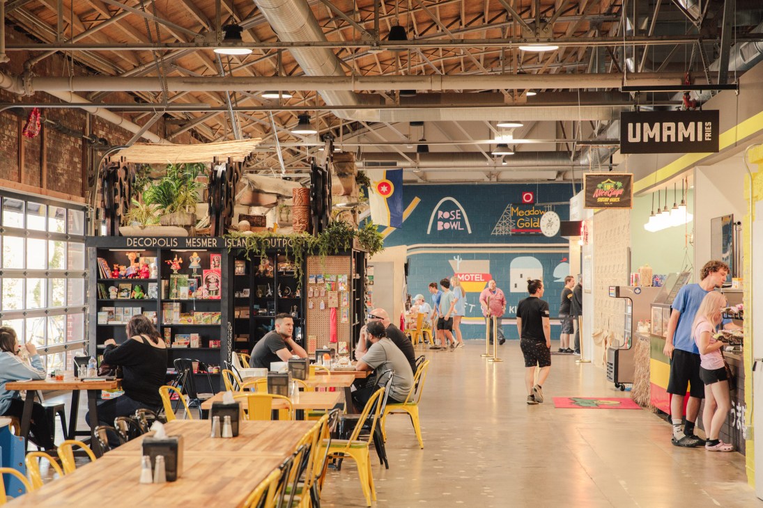 Busy food market interior with people dining at yellow chairs and shopping at vendor stalls during lunch hour.