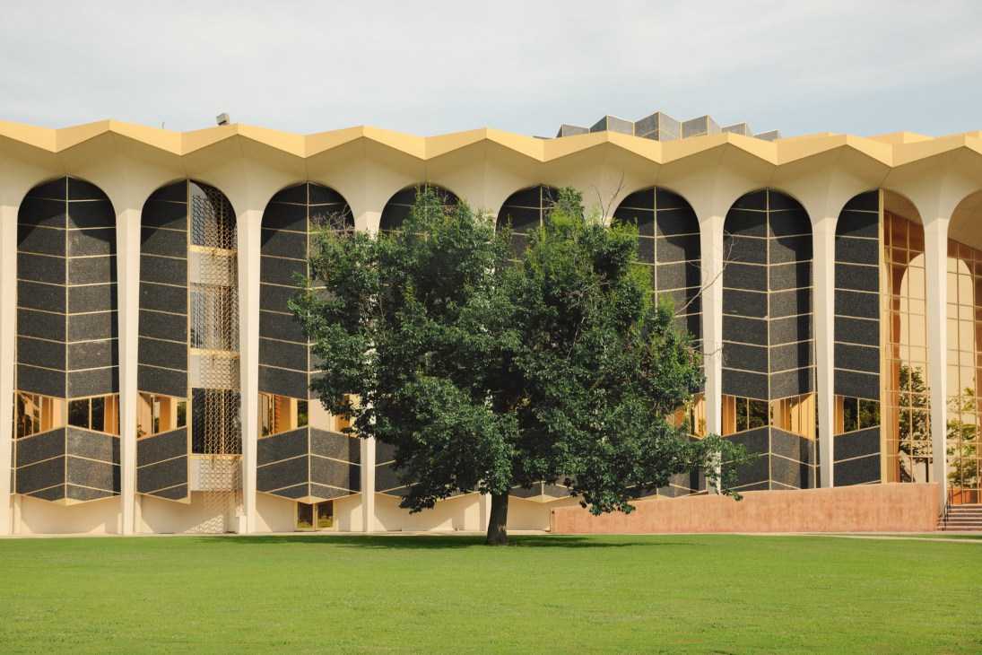 Modern university campus building with distinctive yellow roof, arched windows, and a large tree on green lawn.