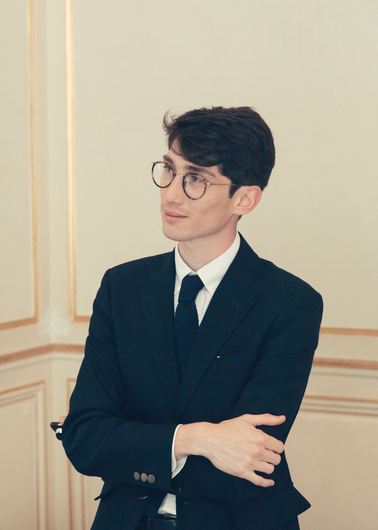 Olivier Flaviano, museum director, wearing round glasses and a dark suit with tie, stands against a pale wall.